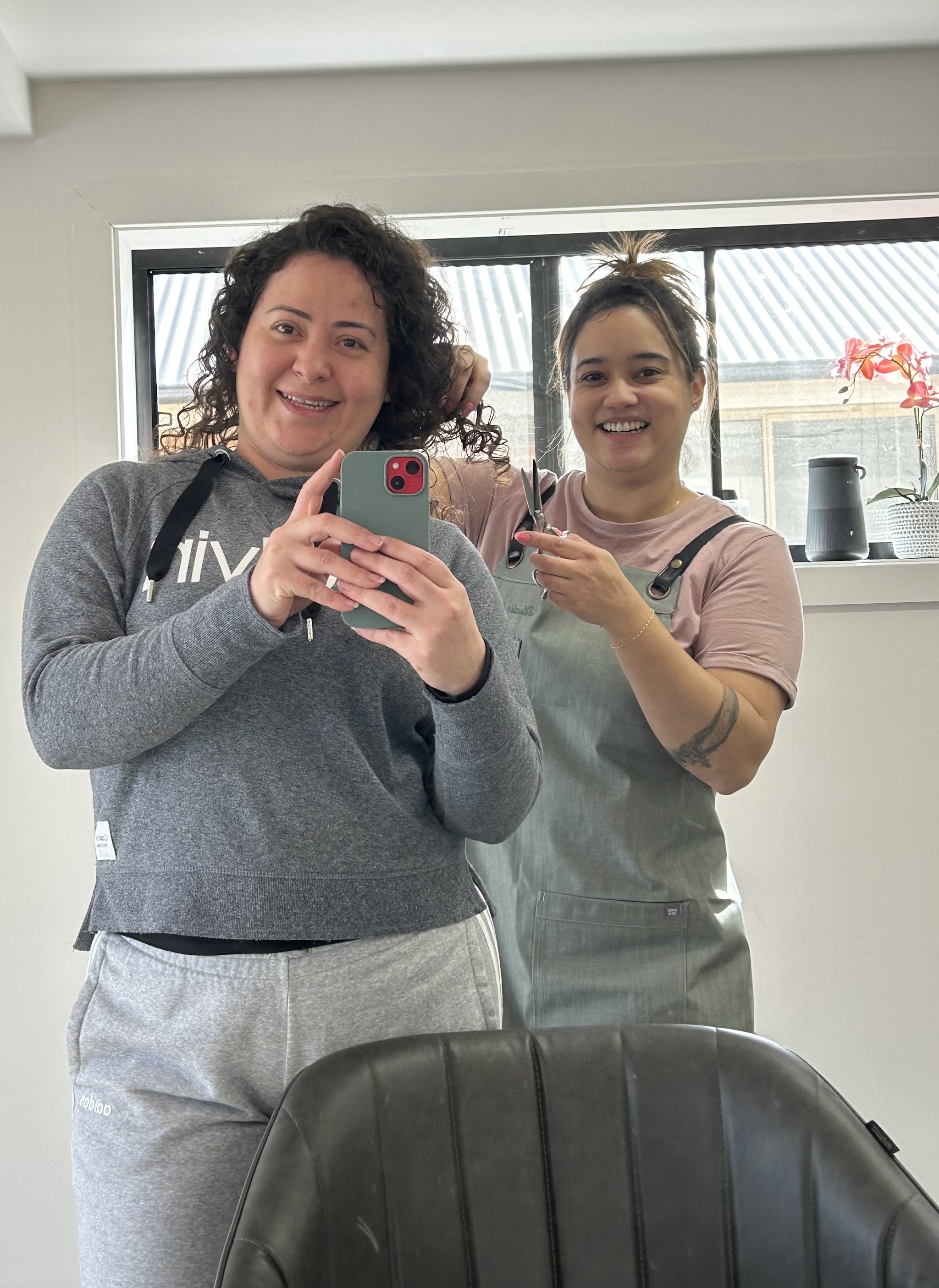 Marilia with scissors mid-appointment, alongside a smiling curly-haired client